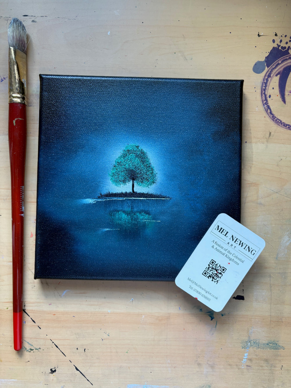 Painting of a green tree on water with a brush and artist's label on a wooden surface
